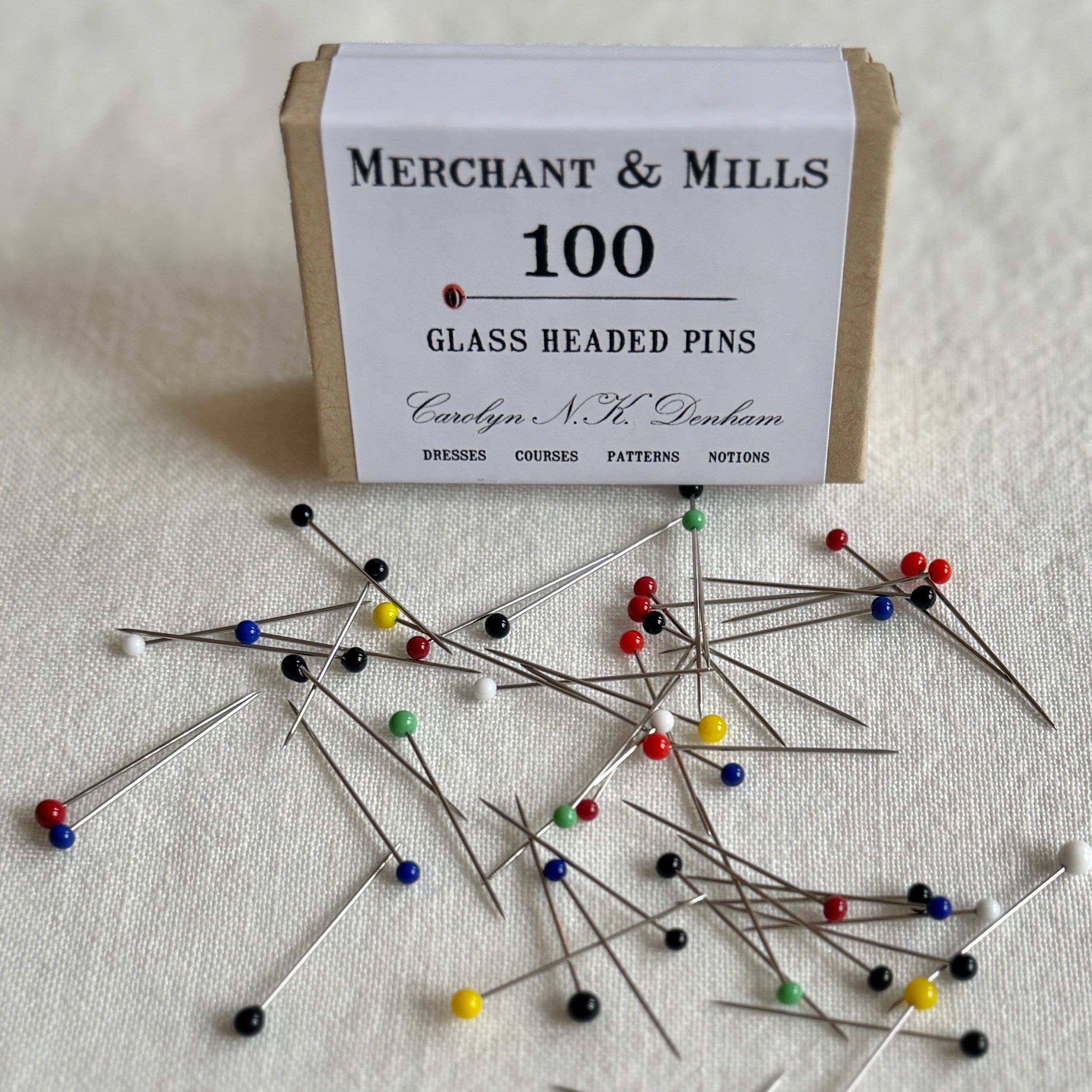 Box of Merchant & Mills glass headed pins with pins scattered on a fabric background